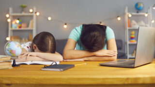 Little girl with her mother lying on table near laptop computer, exhausted from online home schooling. Parent and child tired from making homework