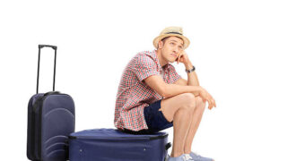 Bored male tourist sitting on his baggage and looking in the distance isolated on white background