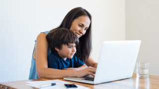 Young mother and son sitting at table and using laptop at home. Smiling mom working at home with her child on the knees while writing an email. Young woman teaching little boy to use the computer.